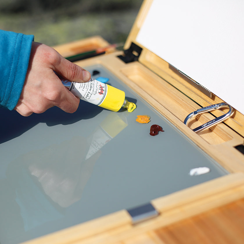 u.go Plein Air Anywhere Pochade Box, 11" x 14.5" model, oil paint being squeezed out on gray tempered glass palette while plein air landscape painting